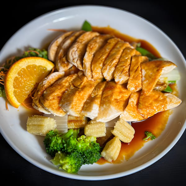 Sliced chicken with orange sauce, broccoli, baby corn, and an orange slice on a white plate.