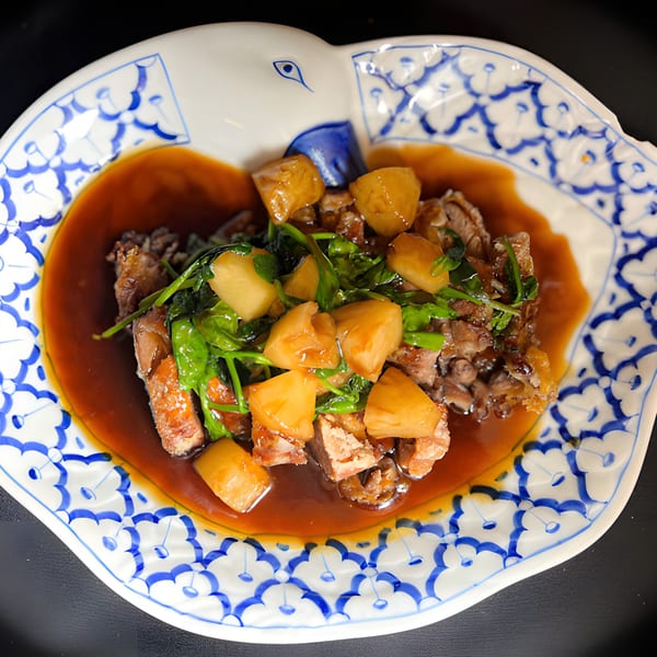 Thai duck dish with pineapple and greens in a rich brown sauce on a decorative white and blue plate.