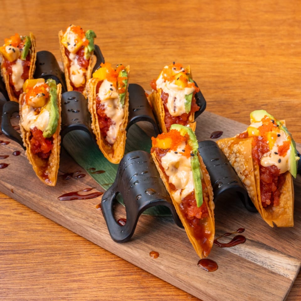 Seven Thai tuna tacos with avocado, mango, and sauce, arranged on a wooden board.