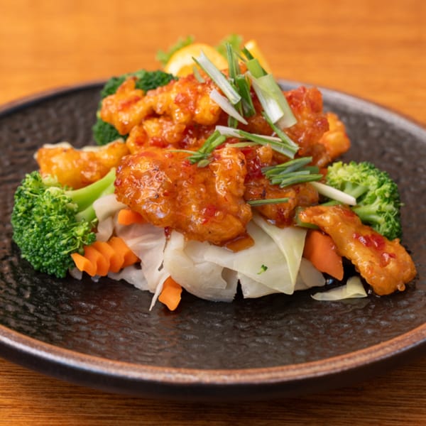 Thai sweet and sour chicken with broccoli, carrots, cabbage, and scallions on a dark plate.
