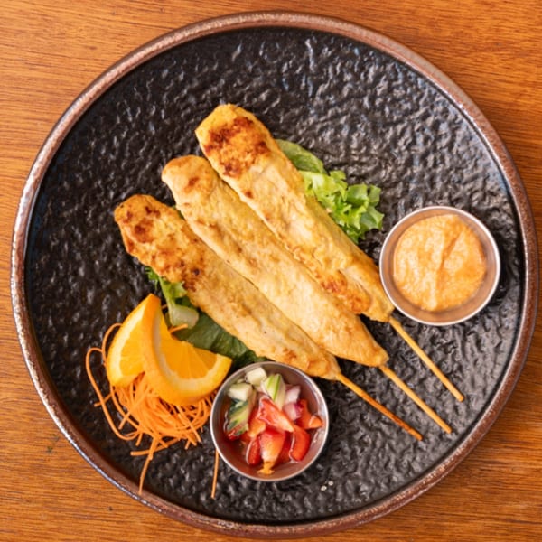 Three chicken satay skewers with peanut sauce, cucumber relish, orange slices, and shredded carrots.