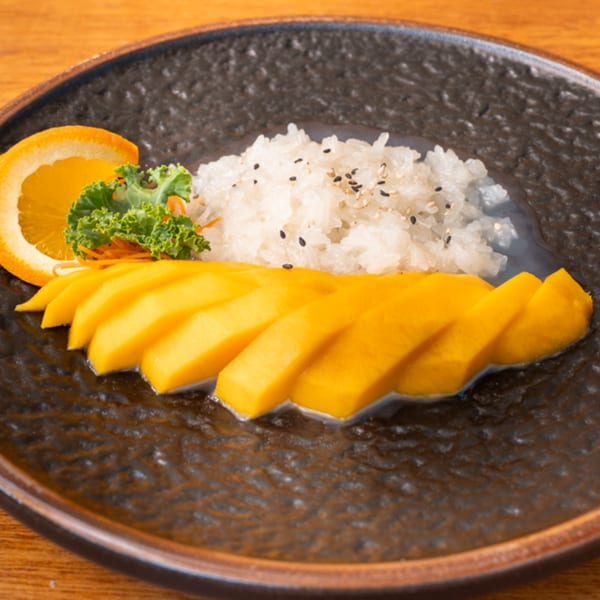 Siam Orchid's mango sticky rice dessert with sesame seeds, an orange slice, and greens on a dark plate.