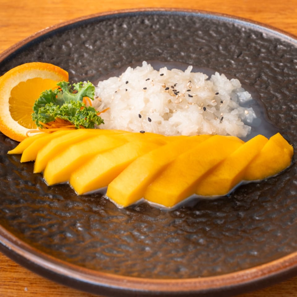 Siam Orchid's mango sticky rice dessert with sesame seeds, an orange slice, and greens on a dark plate.